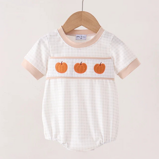 A baby short-sleeve romper in a white and light beige gingham pattern, featuring three orange pumpkin appliqués across the chest. The romper is displayed on a wooden hanger hanging from a light wood clothing rack against a solid, warm off-white background.