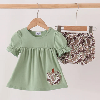 A toddler girl's two-piece outfit hanging on wooden hangers from a wooden rod. The set includes a sage green short-sleeve top with a ruffled neckline and a camouflage-patterned pumpkin applique on the front, paired with matching camouflage-print bloomers.