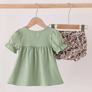 A toddler girl's two-piece outfit hanging on wooden hangers from a wooden rod. The set includes a sage green short-sleeve top with a ruffled yoke and matching sage green puff sleeves, paired with brown and beige camouflage bloomers.