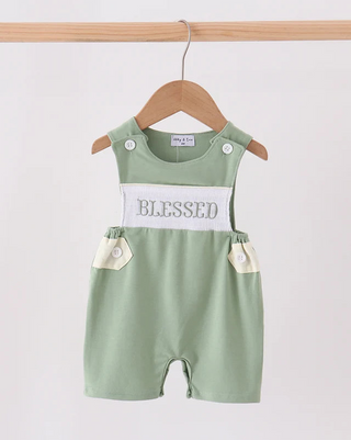 A baby romper in sage green with a white chest panel reading 'BLESSED' and cream-colored pocket flaps, hanging on a wooden hanger against a plain light background.