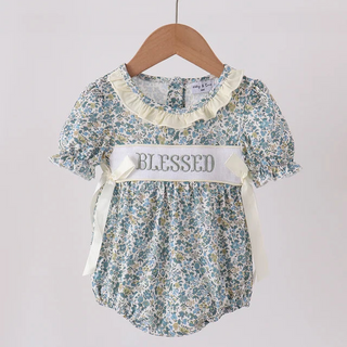 A baby girl's short-sleeve romper with a blue and yellow floral print, featuring a cream ruffled collar, cream ribbon bows, and a white waistband banner that reads 'BLESSED'. The romper is hanging on a wooden hanger from a light wood clothing rack against a plain off-white wall.