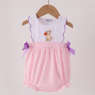 A baby girl's sleeveless romper hanging on a wooden hanger against a light beige wall. The romper features a white top with scalloped edges, a small dog graphic holding a red heart, and a pink and white gingham checkered bottom with purple ribbon bows on the sides.