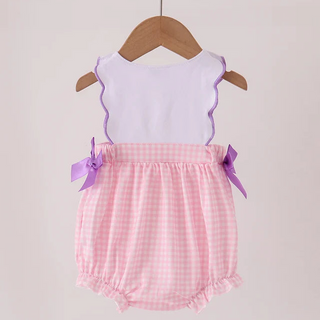 A baby romper hanging on a wooden hanger against a light beige background. The romper has a white sleeveless top with purple ruffle trim, a pink and white gingham checkered skirt, and purple ribbon bows on the sides.