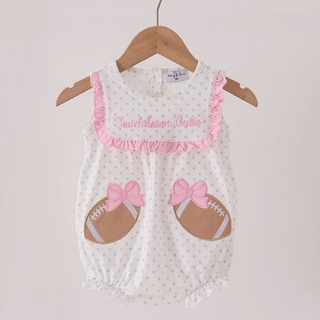 A white sleeveless baby romper with tiny pink polka dots, pink ruffle trim, and two football appliques with pink bows on the front. The romper hangs on a wooden hanger from a wooden clothing rack against a light neutral background.