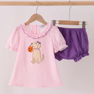 A toddler girl's two-piece outfit hanging on wooden hangers against a plain light background. The set includes a light pink gingham short-sleeve top with a ruffled collar and a cartoon dog holding a pumpkin applique, paired with solid purple bloomers.