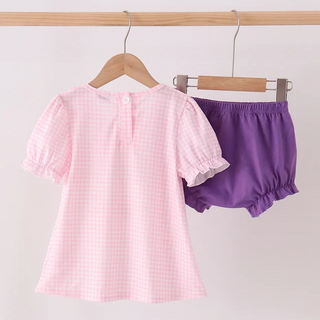 A children's two-piece outfit hanging on wooden hangers from a wooden rod. The set includes a short-sleeve top in light pink and white gingham with ruffled sleeves, and matching purple bloomers.
