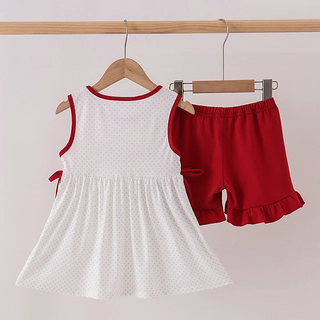 A girl's two-piece outfit hanging on wooden hangers from a wooden clothes rack. The set includes a sleeveless white dress with tiny polka dots and red trim, and matching red ruffled shorts.