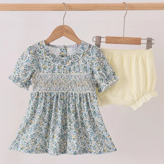 A toddler girl's two-piece outfit hanging on wooden hangers from a wooden rod. The set includes a short-sleeve smocked dress in a light blue and white floral print, and matching smocked bloomers in pale yellow.