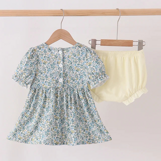 A toddler girl's two-piece outfit set hanging on wooden hangers from a light wood rod. The set includes a short-sleeve dress with a blue, green, and yellow floral print, and a pair of pale yellow ruffled bloomers.