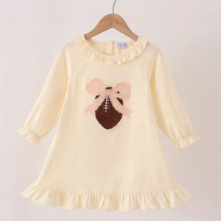 A cream-colored long-sleeve baby dress with a ruffled collar and hem, featuring a football patch with pink bows on the front, hanging on a wooden hanger against a light beige background.