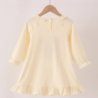A pale yellow long-sleeve baby dress with ruffled trim, hanging on a wooden hanger from a wooden clothes rack against a light beige background.