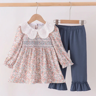A toddler girl's two-piece clothing set hanging on wooden hangers against a plain white wall. The set includes a long-sleeve floral dress with a white lace Peter Pan collar and smocked bodice, paired with dark slate blue pants featuring ruffled hems.