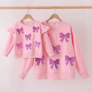 A matching mother-and-child pink sweatshirt set hanging on wooden hangers against a plain light background. The adult sweatshirt is pink with large purple glitter bow prints, and the child's matching sweatshirt has smaller purple glitter bow prints plus ruffled shoulder details.