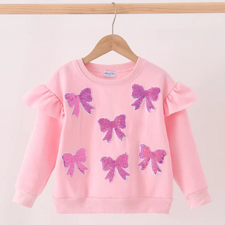 A pink long-sleeve sweatshirt for kids, featuring sequined pink bow designs on the front and ruffled sleeves, hanging on a wooden hanger against a plain light background.