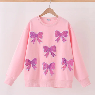 A light pink crewneck sweatshirt hanging on a wooden hanger, decorated with five glittery pink and purple bow appliqués on the front.