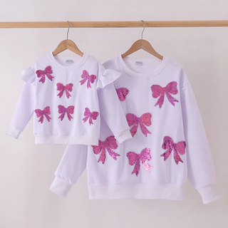 Two white long-sleeve sweatshirts with pink sequined bow designs, displayed on wooden hangers against a plain light background.