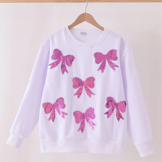 A white crewneck sweatshirt with five pink sequined bow appliqués, hanging on a wooden hanger against a plain off-white wall.