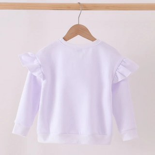 A white  long-sleeve children's sweatshirt with ruffled shoulder details, hanging on a wooden hanger from a wooden clothes rack against a plain off-white wall.