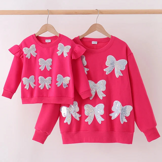 A matching mother and daughter pink sweatshirt set with silver glitter bow appliqués, hanging on wooden hangers against a light pink wall.