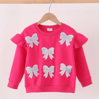 A bright pink long-sleeve children's sweatshirt hanging on a wooden hanger against a light pink background. The sweatshirt features five silver glitter bow appliqués on the front and ruffled details on the shoulders.