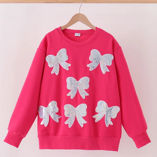 A bright pink crewneck sweatshirt hanging on a wooden hanger, decorated with five large silver glitter bow appliqués on the front, set against a plain light pink background.