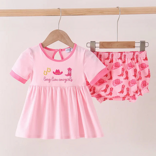 A two-piece baby girl's outfit set featuring a light pink short-sleeve dress with 'Long Legs cowgirls' text and cowboy boot graphics, paired with pink ruffled shorts printed with pink cowboy boots, hanging on wooden hangers.