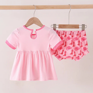A two-piece baby girl's outfit set hanging on wooden hangers from a wooden clothes rack. The set includes a short-sleeve pink dress with a keyhole neckline and matching pink ruffled bloomers featuring a pink and red high-heel print.