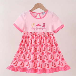 A pink short-sleeve dress hanging on a wooden hanger against a light beige wall. The top portion is solid light pink with 'Long Live cowgirls' and a cowboy hat graphic, while the skirt is patterned with pink cowboy boots and roses.