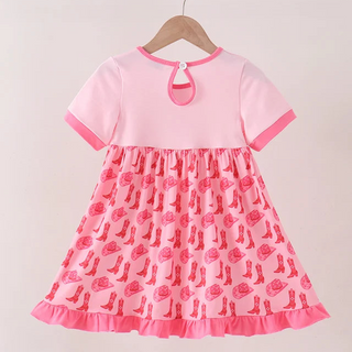 A pink short-sleeve dress with a solid light pink bodice and a pink skirt patterned with cowboy boots and hats, hanging on a wooden hanger against a light beige background.