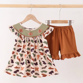 A toddler girl's two-piece outfit hanging on wooden hangers against a plain white wall. The set includes a short-sleeve top with a cream base, an all-over autumn leaf pattern in orange, green, and purple, and a sage green collar with embroidered orange pumpkins.