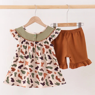 A toddler girl's two-piece outfit set hanging on wooden hangers against a plain white wall. The set includes a short-sleeve dress with a beige base, a green collar, and a multicolor organic spot print, paired with matching rust-brown ruffle-hem shorts.