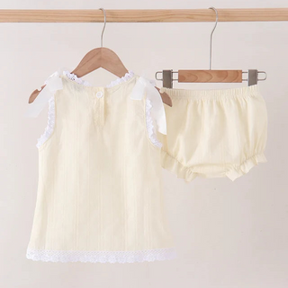 A two-piece baby outfit set consisting of a sleeveless cream top with white lace trim and matching cream bloomers, hanging on wooden hangers from a light wood rod against a plain beige wall.