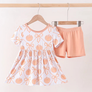 A toddler girl's two-piece outfit set hanging on wooden hangers against a plain light background. The set includes a white short-sleeve dress with an orange pumpkin and bow pattern, and a pair of peach-pink shorts.