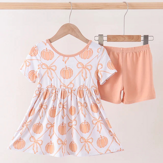 A toddler girl's two-piece outfit set hanging on wooden hangers against a light beige wall. The set includes a short-sleeve white dress with a peach pumpkin and bow pattern, and matching peach shorts.