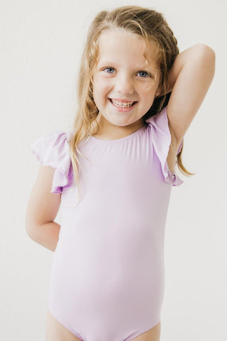 Young girl wearing a light purple leotard with ruffled sleeves on a plain background