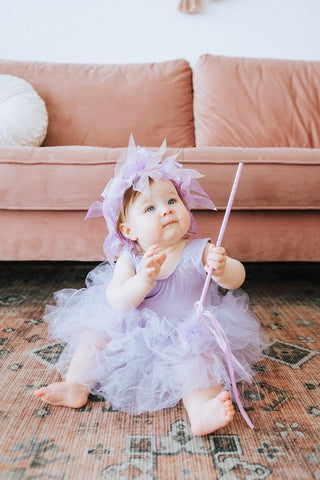 Baby in a purple fairy costume sitting on a patterned rug with a pink couch in the background.