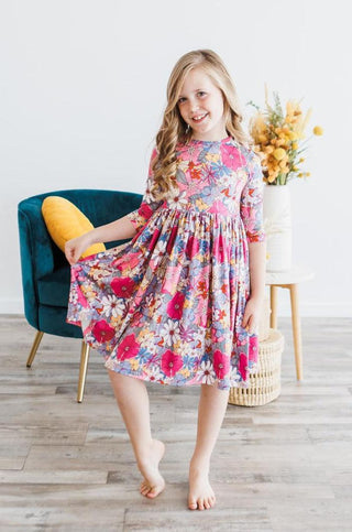 Young girl in a colorful floral dress standing in a room with a chair and flowers.