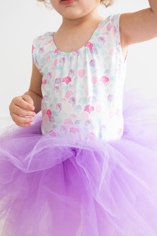 Child wearing a leotard with a mermaid scale print  top and purple tutu skirt on a white background