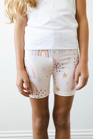Child wearing a white top and patterned shorts with stars and rainbows on a plain background