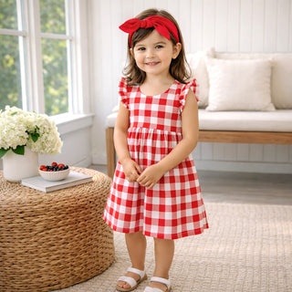 Young girl in a red and white checkered dress standing in a bright room with a couch and flowers.