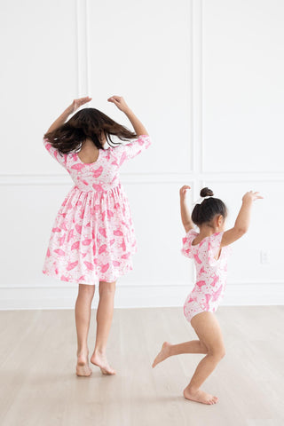 Two children in matching pink ballerina outfits dancing in a room with a white floor and wall.