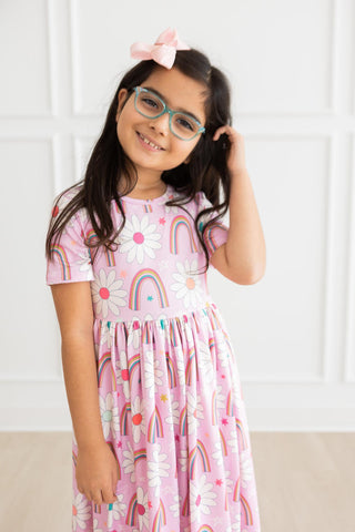 Young girl wearing a pink dress with rainbow and cloud patterns, standing against a white wall.