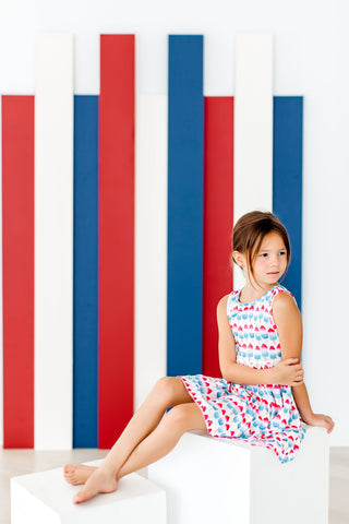 Young girl in a colorful dress sitting on a white surface with red and blue vertical stripes in the background