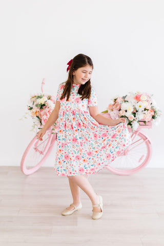 Young girl in a floral dress standing next to a pink bicycle with flower baskets on a white background
