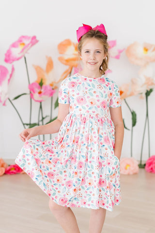 Young girl in a floral dress with pink flowers and a pink bow, standing in front of a floral backdrop.