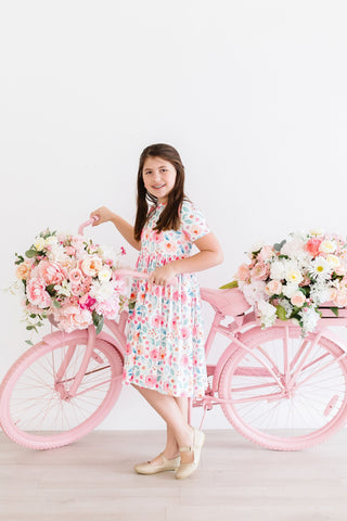 Young girl in a floral dress standing next to a pink bicycle with flower baskets on a white background