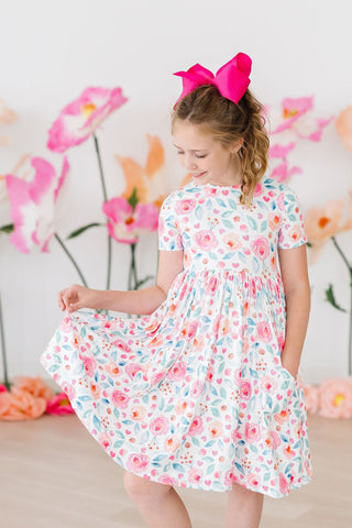 Young girl in a floral dress with a pink bow in her hair, standing against a floral backdrop.