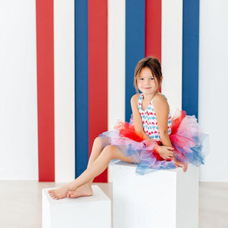Young girl in a colorful leotard sitting on a white block with red and blue striped background