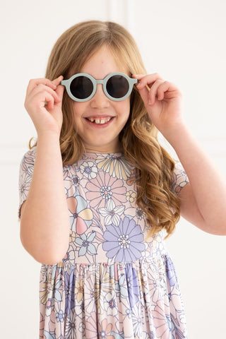 Young girl wearing a floral dress and holding sunglasses up to her face against a white background