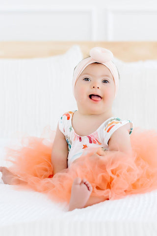 Baby in a colorful leotard with a peach-colored tutu sitting on a white surface.
